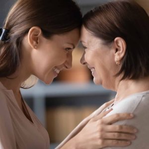 Smiling middle-aged mom and adult daughter touch foreheads look in eyes sharing close intimate moment together, happy senior mother and grownup girl child hug show love and understanding at home